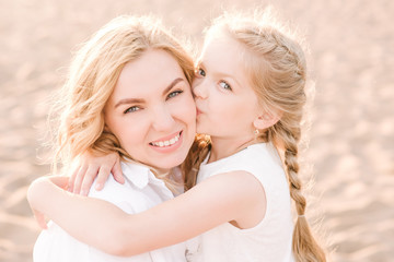 A girl hugs and kisses her mother on the cheek. Mom’s daughter are on the beach, dressed in white clothes. The concept of love between mom and daughter. 