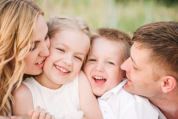 A family of four dressed in bright clothes. Dad and mom hug daughter and son, kiss them on the cheeks. The kids are laughing. The concept of a happy family.