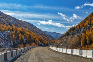 Gravel road with white and black fence in Siberian mountains with snow and yellow trees