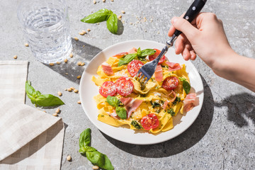 partial view of woman eating tasty Pappardelle with tomatoes, pesto and prosciutto near water on grey surface