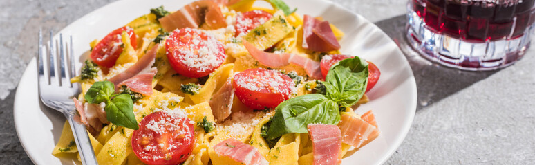 tasty Pappardelle with tomatoes, pesto and prosciutto with fork near red wine on grey surface, panoramic shot