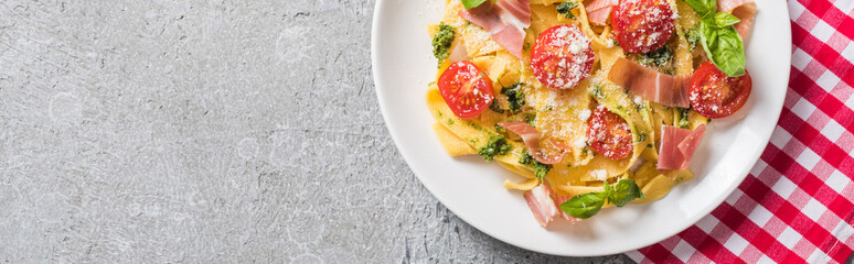 top view of cooked Pappardelle with tomatoes, basil and prosciutto on plaid napkin on grey surface, panoramic shot