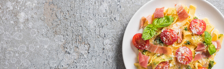 top view of cooked Pappardelle with tomatoes, pesto sauce and prosciutto on grey surface, panoramic shot