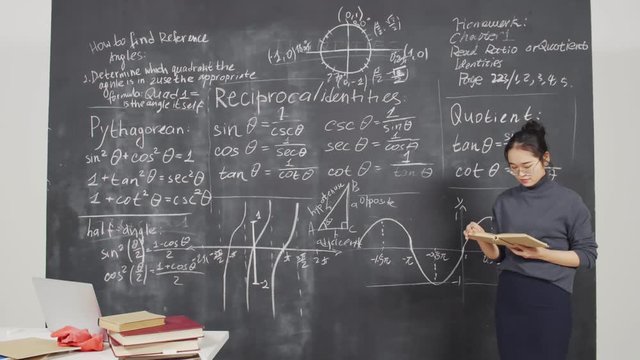 Lockdown of young Asian woman standing with book in her hands, analyzing it and then looking at blackboard and comparing information