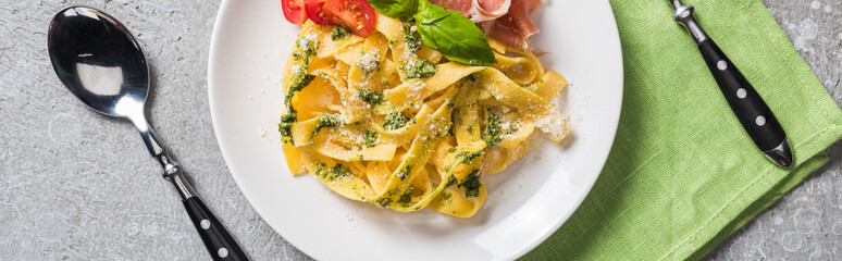 top view of cooked Pappardelle with tomatoes, basil and prosciutto near cutlery on grey surface, panoramic shot