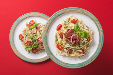 top view of cooked Pappardelle with tomatoes, basil and prosciutto on plates on red background
