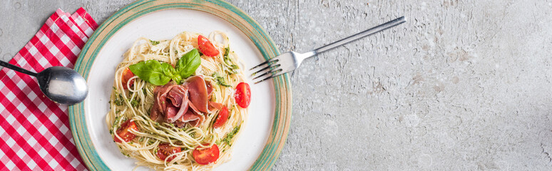 top view of served Pappardelle with tomatoes, basil and prosciutto on plaid napkin with cutlery on grey surface, panoramic shot