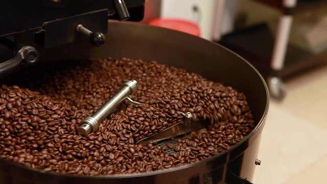 Person's hand uses a small clear scoop to push freshly roasted coffee beans as they are moved and mixed in roaster's cooling tray.