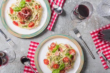 top view of Pappardelle with tomatoes, basil and prosciutto on plates on plaid napkins with cutlery near red wine and water on grey surface