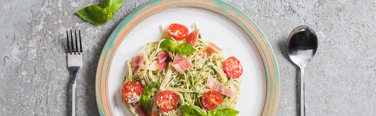 top view of cooked Pappardelle with tomatoes, basil and prosciutto near cutlery on grey surface, panoramic shot