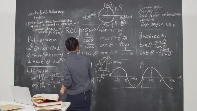 Lockdown of young female Asian woman standing at blackboard, taking rag, wiping blackboard then taking piece of chalk, making corrections on blackboard, taking book and starting reading it
