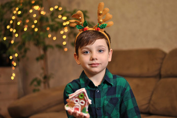 Cute little kid boy holds toy house wearing Christmas deer horns dressed up in green shirt with garland lights bokeh. Xmas winter holiday children lifestyle portrait. Beautiful New Year people
