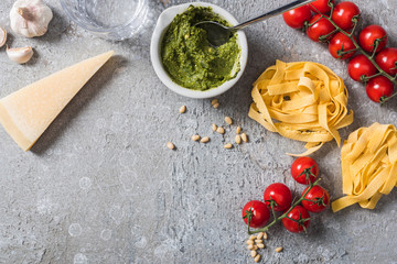 top view of raw Pappardelle near tomatoes, garlic, pine nuts, water and pesto sauce on grey surface