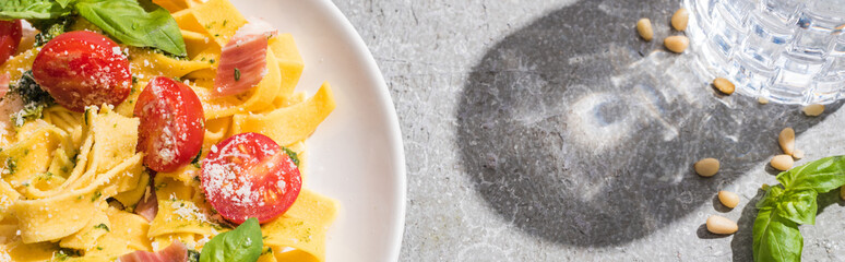 top view of tasty Pappardelle with tomatoes, pesto and prosciutto near water on grey surface, panoramic shot