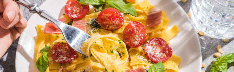 partial view of woman eating tasty Pappardelle with tomatoes, pesto and prosciutto near water on grey surface, panoramic shot
