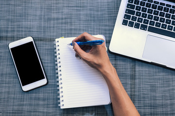 Top view of a right hand of a woman writing on a notebook. Phone with blank and keyboard of a laptop