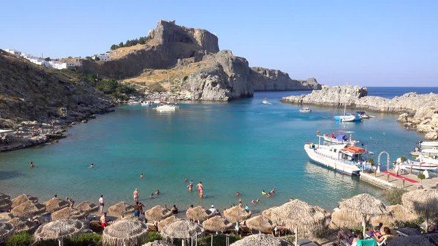 St. Paul's beach, Bay, Mediterranean sea, people swimming, sun umbrellas, boats, fortress in the background, Lindos city Rhodes island Greece. September 2019