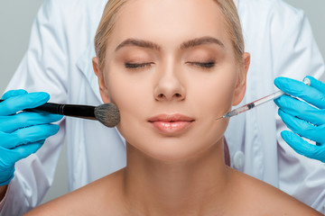 cropped view of beautician in latex gloves holding cosmetic brush and syringe near attractive girl with closed eyes