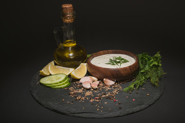 Bowl with tzatziki sauce with ingredients, spices and olive oil on dark board on black background