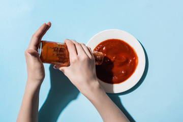 Cropped view of woman pouring tomato sauce from bottle to plate on blue background