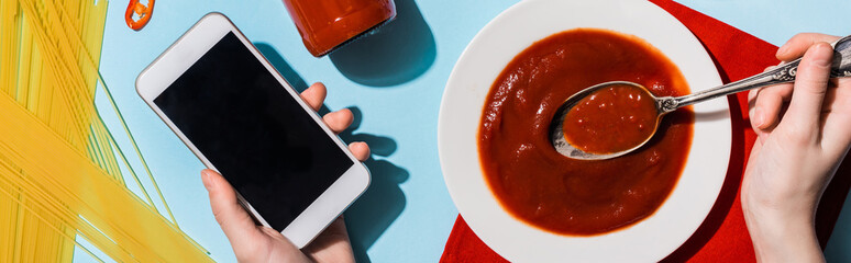 Cropped view of woman holding smartphone beside ketchup and spaghetti on blue background, panoramic shot