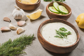Homemade tzatziki sauce in wooden bowl with ingredients and greenery on stone background