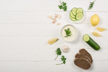 Top view of tzatziki sauce ingredients, greens and bread on white wooden background