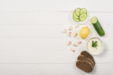 Fototapeta na wymiar Top view of tzatziki sauce with organic ingredients and bread on white wooden background