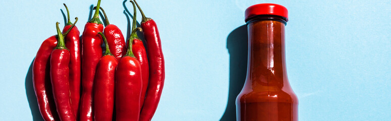 Top view of chili sauce in bottle with chili peppers on blue background, panoramic shot
