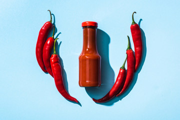 Top view of chili sauce in bottle between chili peppers on blue background