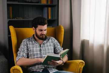 Serious young man with beard is sitting on yellow chair and reading book. Concept of everyday life.