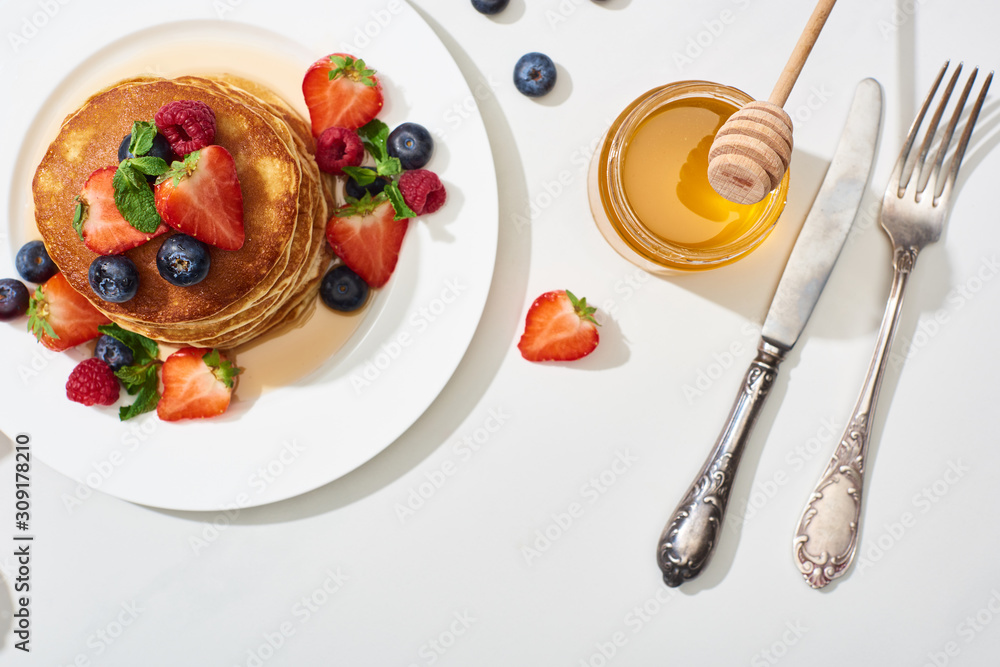 Wall mural top view of delicious pancakes with honey, blueberries and strawberries on plate near cutlery on marble white surface