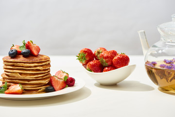delicious pancakes with blueberries and strawberries on plate near herbal tea in teapot on white surface isolated on grey