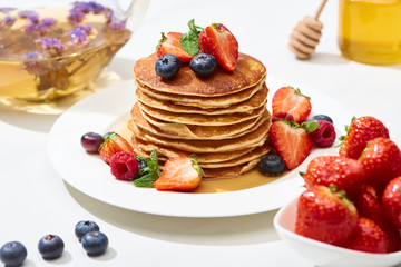 selective focus of delicious pancakes with honey, blueberries and strawberries on plate on white surface