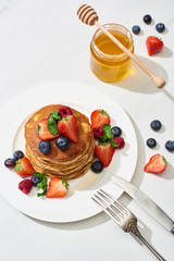 top view of delicious pancakes with honey, blueberries and strawberries on plate near fork and knife on marble white surface