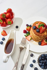 top view of delicious pancakes with maple syrup, blueberries and strawberries on plate near fork and knife on marble white surface