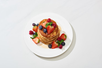 top view of delicious pancakes with honey, blueberries and strawberries on plate on marble white surface