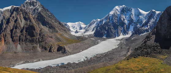 A large glacier lies in a mountain valley. Rocky steep slopes, snow-capped peaks. Panoramic mountain view, Altai.