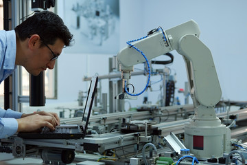 Focus on robot arm which picks up product from automated car on production line and an engineer working on laptop to programming. Industry 4.0 concept; artificial intelligence in smart factory.