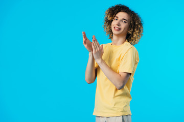 smiling curly teenager applauding isolated on blue