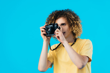 curly teenager taking picture on film camera isolated on blue