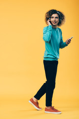 smiling curly teenager holding smartphone and listening music in headphones while walking on yellow