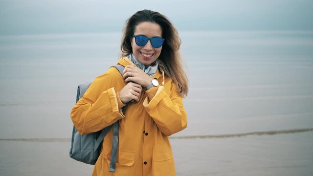 Smiling lady in a yellow coat is wearing sunglasses near sea and cloudy sky on background.