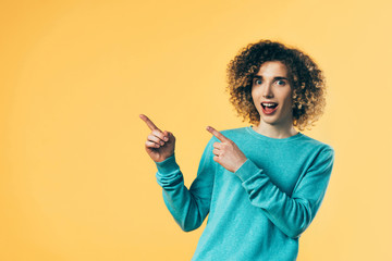 curly teenager with open mouth pointing with fingers aside isolated on yellow