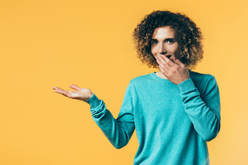 smiling curly teenager covering mouth and pointing with hand aside isolated on yellow