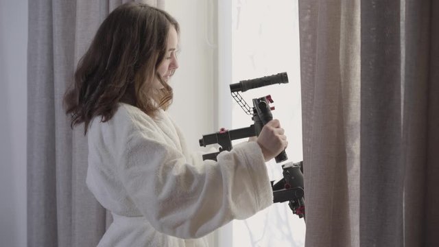 Side view portrait of young Caucasian brunette woman standing at the window with video equipment. Beautiful female videographer holding stabilizer in the morning. Profession, creativity, lifestyle.
