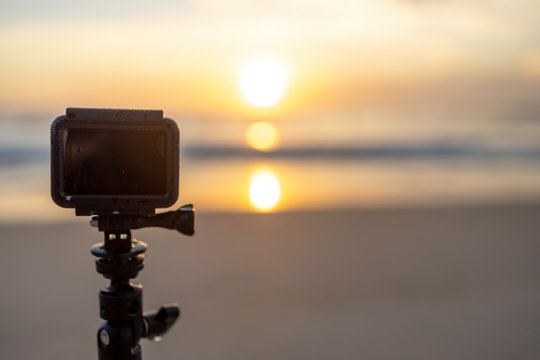 Small action camera on a tripod stand on a beach in Rio taping and watching the sunrise with the sun reflecting in the ocean water