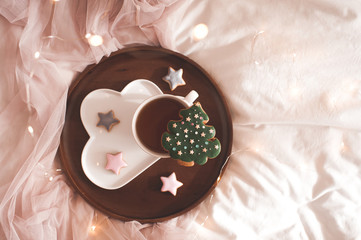 Tasty ginger bread in Christmas tree shape staying on cup of tea on wooden tray in bed over glowing lights closeup. Top view. Xmas. Winter holiday season.