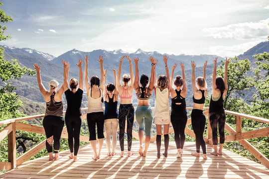 Rear view of group of joyful happy athletic young women in gymnastic clothes raise their hands up and enjoy joint activities in open air among mountains. Concept of sports and personal achievements