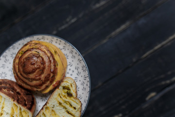 One whole and two halfs of cheese and dill bread bun on plate standing on black wooden background. Tasty unhealthy food. Diet, nutrition, eating habits concept. Place for text. 